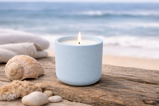 Lit light-blue candle in a cast-stone vessel on a weathered wooden surface by the beach, with seashells and stones in the foreground and blurred ocean waves in the background.