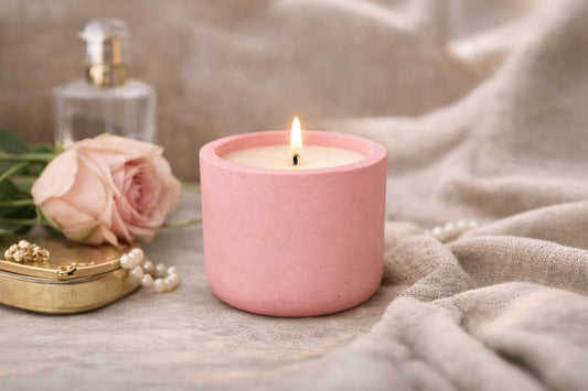 Lit blush-pink candle in a cast-stone vessel, with a pink rose, perfume bottle, and gold jewellery tray in the background.